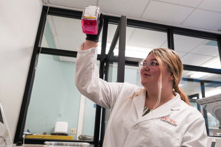 A student wearing a white lab coat holds up a beaker