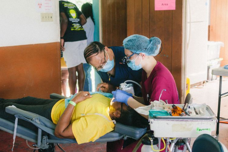 Two volunteers wearing caps and masks work on a patient wearing a yellow shirt lying down in a dental chair